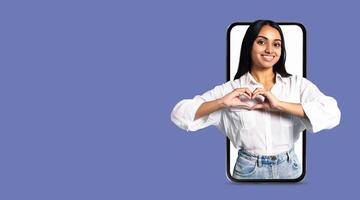 A young woman smiles and creates a heart shape with her hands, standing next to a large smartphone displaying her gesture. This scene symbolizes communication and connection through technology. photo