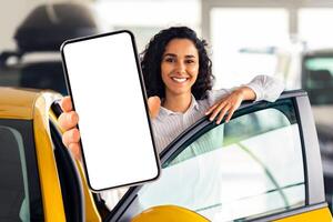 Positive millennial middle eastern woman standing by brand new yellow car, showing smartphone with empty screen and smiling at camera, mockup, renting auto on Internet. Car dealership online photo