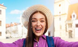 A joyful woman in a straw hat takes a selfie while standing in an urban area. The sun shines on her face as she engages with facial recognition technology. photo