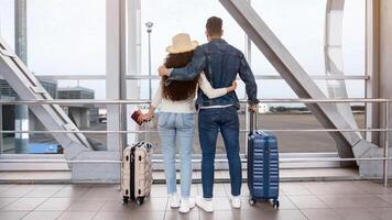 Rear View Of Romantic Couple Looking Out Of Window At Airport Terminal, Unrecognizable Spouses Ready For Summer Vacation, Standing With Luggage And Embracing, Enjoying Travelling Together, Copy Space photo