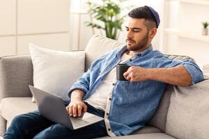 Portrait of focused handsome young jewish guy wearing yarmulke using personal computer holding mug and drinking coffee or tea. Man sitting on sofa and working or watching , movie or webinar photo