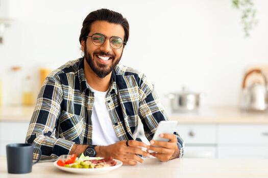 alegre Medio este joven hombre con largo pelo y elegante barba en casual disfrutando desayuno, Bebiendo café, participación teléfono inteligente, indio chico persona de libre dedicación comprobación correos electrónicos en el mañana, Copiar espacio foto