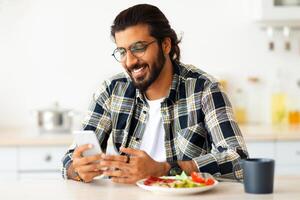 Cheerful bearded long-haired middle-eastern guy with glasses enjoying lunch at home, sitting at kitchen table alone, using mobile phone and smiling, chatting with girlfriend, copy space photo