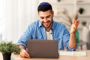 Virtual Meeting Concept. Portrait of cheerful israeli man in kippah having making online conference with colleagues or employees, sitting at table at home office and explaining, working remotely photo
