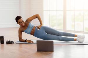 Full Body Length Of Happy Fit African American Woman Exercising On Yoga Mat Standing In Side Plank Position Near Window, Looking At Computer Screen Watching Tutorial Or Virtual Workout With Trainer photo