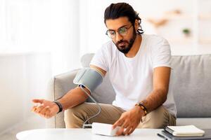 Young arab man sitting on couch, checking blood pressure and taking notes at home. Indian guy in white t-shirt using modern tonometer, experiencing hypertension caused by stress, copy space photo