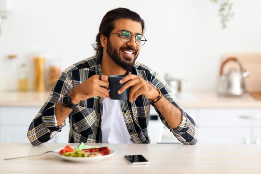 sonriente árabe chico en casual sentado a cocina escritorio a hogar, participación jarra con café, teniendo desayuno solo, mirando a Copiar espacio. sano dieta y estilo de vida para millennials concepto foto