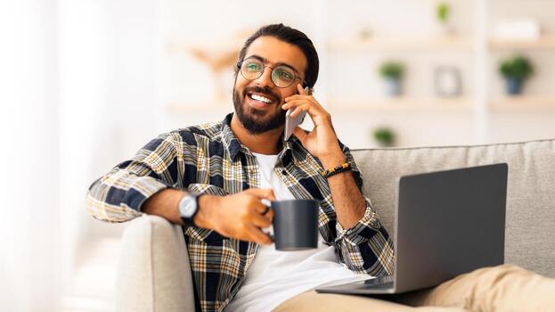 relajado árabe chico sentado en sofá con ordenador portátil y jarra con caliente beber, teniendo teléfono cobertura mientras Relajado a hogar, mirando a Copiar espacio y sonriente. Doméstico entretenimiento y tecnologías foto
