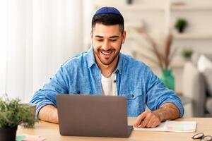 Telecommuting Concept. Potrait of smiling casual israeli man sitting at desk, using laptop. Male watching webinar looking at pc screen, working or studying remotely, free copy space, banner photo