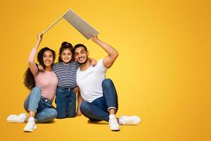 Real Estate Concept. Smiling Arab Parents With Little Daughter Holding Symbolic Carton Roof Above Their Heads, Happy Middle Eastern Family Dreaming Of New Home While Sitting Over Yellow Background photo
