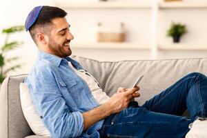 Mobile Communication. Side view of happy young jewish man using modern smartphone device while sitting on sofa at home, modern design interior. Cheerful guy typing an sms message at social network photo
