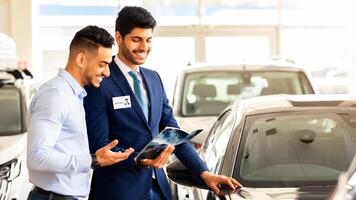 Handsome young arab man having conversation with sales assistant in showroom, side view, copy space. Cheerful car dealer showing catalogue with luxury autos, selling nice automobiles in new salon photo