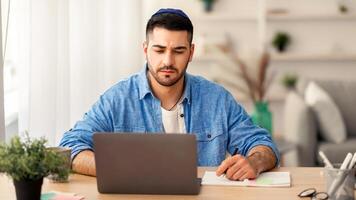 E-learning And Online Training. Portrait of focused jewish man in kipa working or studying remotely at home office, writing in his notebook with pen, taking notes, using laptop looking at screen photo