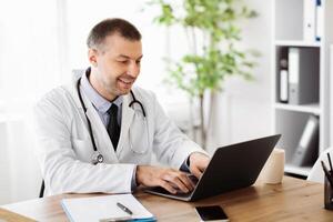 Doing Research Concept. Portrait of smiling middle aged specialist physician using computer laptop, typing on keyboard, sitting at table in hospital, giving online consulation, telemedicine photo
