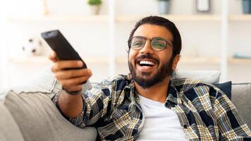 Relaxed arab guy wathcing movie on TV at home, sitting on couch alone, having happy face expression. Happy young man with TV remote watching comedy and laughing, copy space, closeup photo