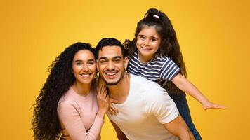 Family Time. Portrait Of Happy Arab Parents And Daughter Having Fun Together While Posing Over Yellow Background In Studio, Positive Middle Eastern Mom, Dad And Child Smiling At Camera, Copy Space photo