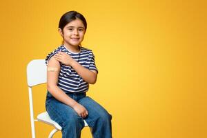 Portrait Of Cute Little Arab Girl Showing Arm After Coronavirus Vaccination, Adorable Middle Eastern Child Got Covid-19 Vaccine Injection, Sitting With Rolled Up Sleeve Over Yellow Background photo