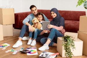 Modern Remote Communication Concept. Portrait of positive arabic family making virtual call with friends or grandparents using tablet, sitting on floor carpet with paint, talking online photo