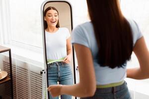 Weight Loss. Happy Girl Measuring Waist With Tape Standing In Front Of Mirror Slimming At Home. Copy Space, Selective Focus photo