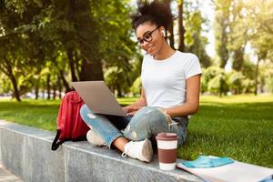 Modern Education. Black Girl Using Laptop Computer Learning Online Having Lesson Via Call Outdoors. Happy Student Watching Virtual Lecture Or Webinar In Park. Side-View Shot photo