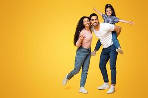 Cheerful Middle Eastern Family Of Three Having Fun Together Over Yellow Background, Happy Young Arab Parents And Their Little Daughter Laughing And Smiling At Camera, Full Length Shot, Copy Space photo