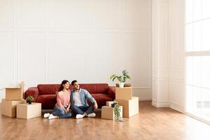 Moving House. Cheerful Middle Eastern Spouses Sitting Among Unpacked Moving Boxes In New Home, Hugging And Looking Aside At Window. Real Estate Ownership And Apartment Rent Concept. Free Copy Space photo