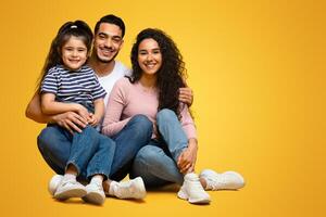 Happy middle eastern family sitting on floor and embracing, cheerful loving arab parents posing together with their cute little daughter over yellow studio background, looking at camera, copy space photo