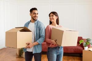 Loving couple carrying cardboard boxes together, standing back to back looking at each other in living room on moving day. Young tenants relocating to new apartment with belongings, unpacking things photo