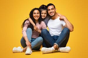Portrait Of Young Happy Middle Eastern Family With Little Daughter Posing Together, Cheerful Arab Parents And Their Child Embracing While Sitting On Floor Over Yellow Background In Studio, Free Space photo