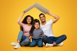 Family Housing And Insurance. Happy Arab Family With Little Daughter Sitting Under Symbolic Carton Roof, Young Middle Dreaming Of New Home While Posing Together Over Yellow Background In Studio photo
