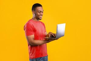 Happy African American Freelancer Man Using Laptop Standing In Studio Over Yellow Background. Black Guy Browsing Internet Holding Computer. E-Learning, Technologies Concept. Side View photo
