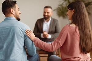 Marital Therapy Concept. Happy couple reconciling at session in therapists office during appointment, looking at each other, smiling woman touching man's shoulder, back view, selective focus photo