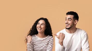 Joyful Arab Man And Woman Looking Aside And Pointing At Copy Space With Thumbs Up, Interested Middle Eastern Couple Sitting On Floor Over Yellow Studio Background And Showing Free Place For Ad photo