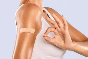 Effective Vaccine Against Covid. Close Up Cropped View Of Female Patient Showing Vaccinated Arm With Sticking Patch On Her Shoulder After Getting Shot In Clinic And Okay Sign Gesture, Blue Studio photo