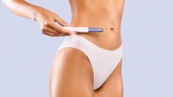 New Life Concept. Closeup cropped view of unrecognizable pregnant woman in white panties holding positive pregnancy test with 2 two red lines standing isolated on blue studio background, banner photo
