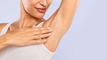Closeup cropped view of happy woman smelling her fresh armpit isolated on blue studuo background, putting hand up, touching axilla. Free copy space, advertisement for under arm deodorant or epilation photo