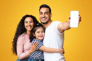 Cheerful Arab Family Of Three Demonstrating Blank Smartphone With White Screen, Loving Middle Eastern Mom, Dad And Little Daughter Embracing And Enjoying New Mobile Application Or Website, Mockup photo