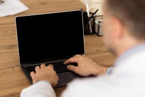 Lifestyle And Gadgets Concept. Over the shouder view of adult man using laptop with blank black screen for mockup template, sitting at wooden table, back shot. Person typing on computer keyboard photo