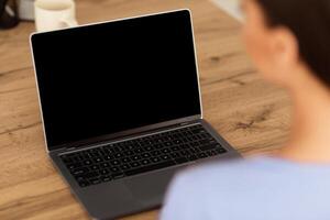 Lifestyle And Modern Technology Concept. Woman using laptop with blank black screen for mock up template, sitting at wooden desk, view over the shoulder, back shot. Person working on notebook photo