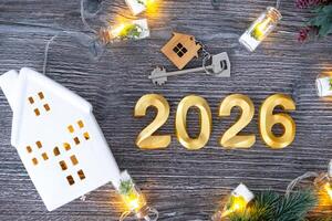 New Year 2026 golden numbers House key tiny form of home on festive grey wooden background flatlay, lights of garlands, greeting card. Housing, real estate, relocation, mortgage, insurance photo