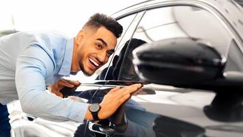 Handsome young arab man buying new car, standing at car salon and checking automobile surface, closeup. Smiling middle-eastern guy looking at beautiful black auto in showroom photo