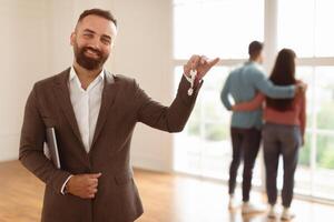 House Ownership. Portrait Of Smiling Professional Real Estate Agent In Suit Holding And Showing Key To Camera, Selective Focus. Happy Spouses Choosing New Home Looking Out Window In Blurred Background photo