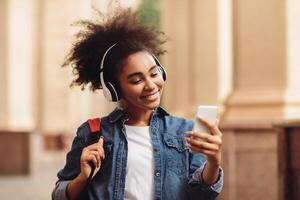 Educational Podcast. African American Student Girl Using Phone Wearing Wireless Headphones Walking At University Campus Outdoor. People And Gadgets, E-Learning Application Concept photo