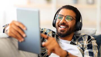 Happy bearded middle-eastern man wearing glasses using entertaining mobile application on pad and headphones while relaxing on couch at home, joyful indian guy looking for movie online, holding tablet photo