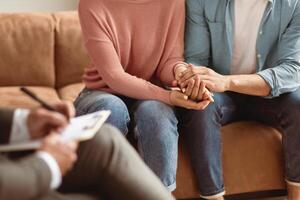 Help Concept. Closeup Cropped View Of Spouses Having Session With Man Marriage Therapist, Sitting On Couch In Office And Holding Each Other Hands. Consultant Writing Taking Notes, Selective Focus photo