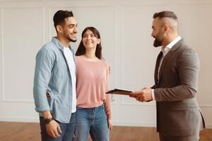 Portrait Of Smiling Professional Man Realtor In Suit Showing New Apartment To Young Arab Couple, Holding Clipboard. Happy Middle Eastern Guy And Lady Hugging And Talking With Seller photo