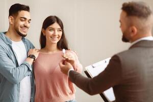 Happy smiling Middle Eastern couple taking key of future new apartment. Professional real estate agent wearing suit holding in hand and giving keys to young buyers. Mortgage, Property For Sale Concept photo
