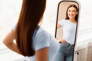 Smiling Girl Looking At Mirror After Slimming Happy About Dieting Result Standing Indoors. Weight Loss. Selective Focus photo