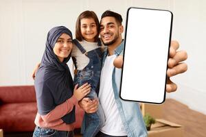 Portrait Of Happy Muslim Family Showing Keys, Cheerful Parents Hugging Little Kid, Holding Daughter On Hands. Arabic People Moving In New Apartment Standing In Living Room. Selective Focus photo