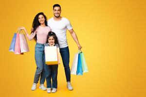 Family Shopping. Happy Young Middle Eastern Parents And Their Cute Little Daughter Holding Shopper Bags While Posing Together Over Yellow Background In Studio, Enjoying Seasonal Sales, Copy Space photo